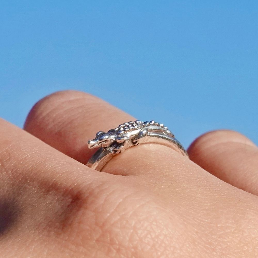 A silver ring shaped like a small alligator or crocodile, worn on a person's finger against a bright blue sky background.